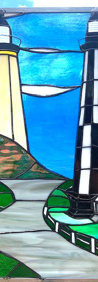 A large vertical rectangular stained glass panel showing two lighthouses and a path leading through the middle of the image