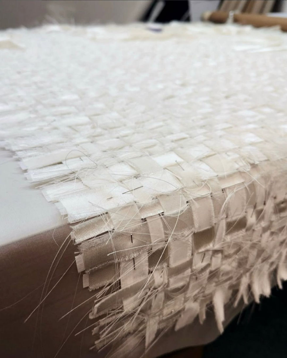 Close-up of an unfinished white silk weave made from overlapping fabric strips, with loose threads and pins visible along its edge.