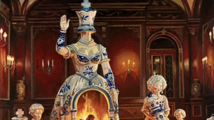 A porcelain family stands among chess pieces, led by a tall faceless figure with a lit fireplace built into its torso.