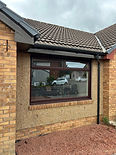 A newly installed window with white frames on a grey house, surrounded by discarded wooden planks and a red fence in the background.