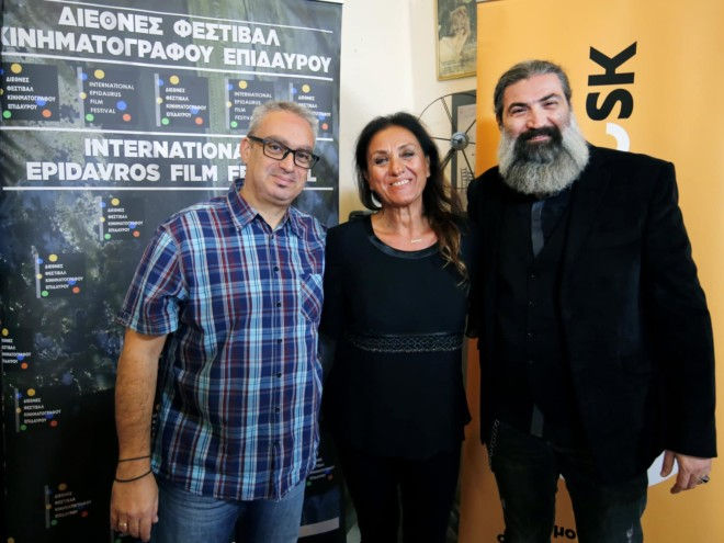Three people smile in front of banners for the Epidaurus Film Festival. One wears a plaid shirt, one in black; setting is cheerful, casual.