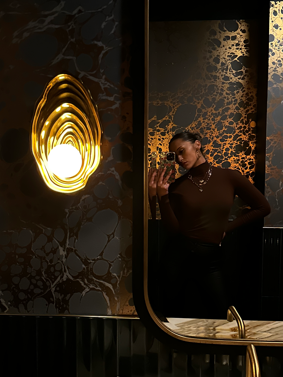 Giovanna taking a mirror selfie in a dimly lit bathroom with ornate gold and black wallpaper. Elegant mood with stylish attire and decor.