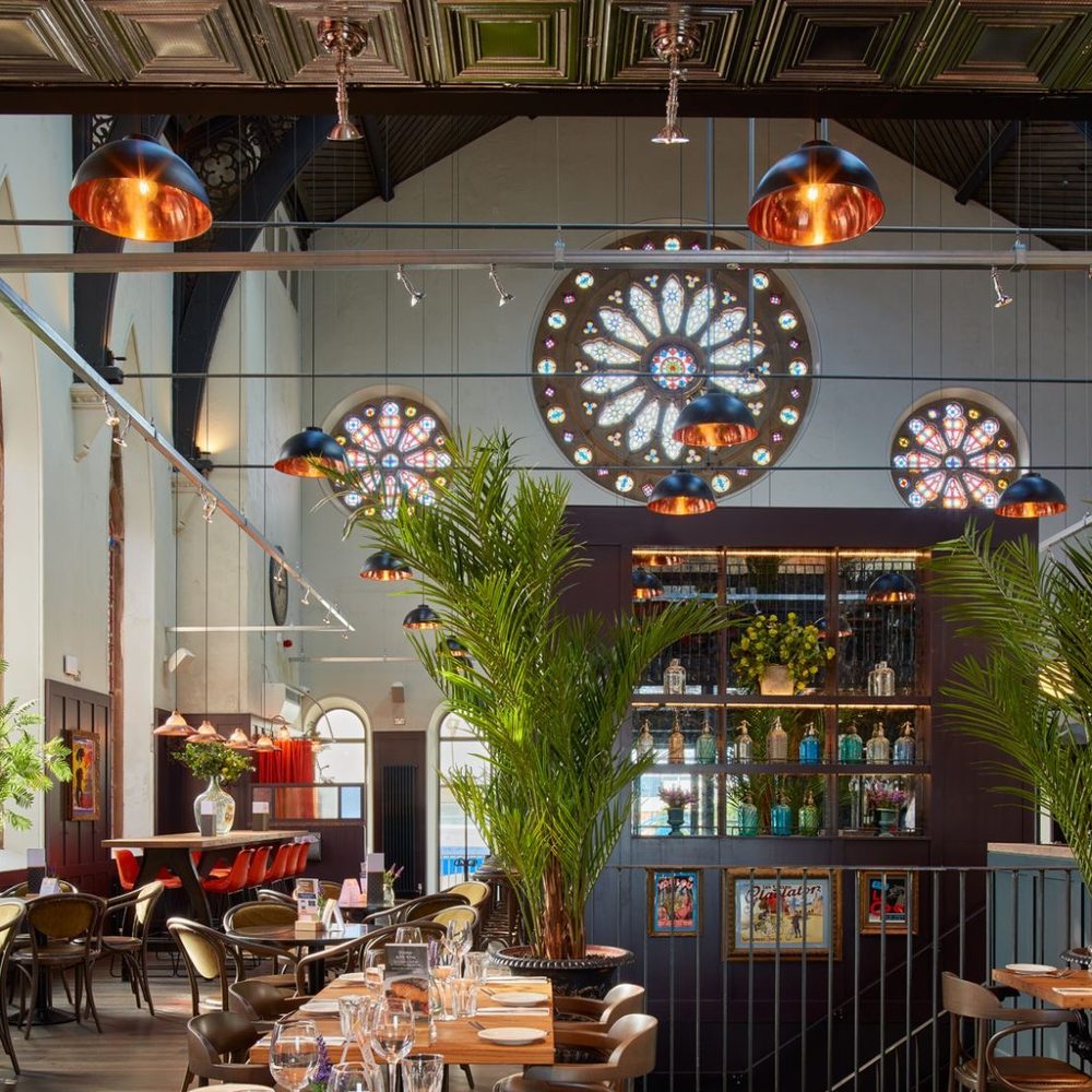 Restaurant in a converted church with stained-glass rose windows, copper pendant lights, palms and tables set for dinner