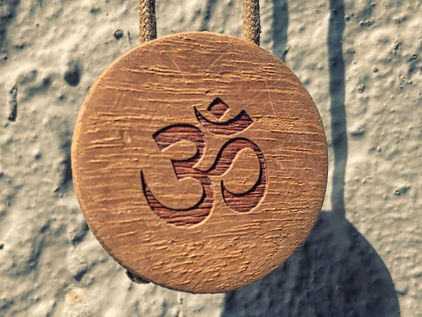 Holzanhänger mit eingraviertem AUM-Symbol hängt an rauer, hellgrauer Wand. Warme Farbtöne, länglicher Schatten sichtbar. Ruhige Atmosphäre.