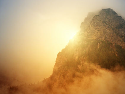 Ein majestätischer Berggipfel, der teilweise von weichem, goldenem Nebel umhüllt ist. Die Sonne geht hinter dem Berg unter oder steigt auf und wirft ein warmes, leuchtendes Licht. Der Vordergrund zeigt neblige Wolken, die ein Gefühl von Geheimnis und Ruhe hinzufügen.