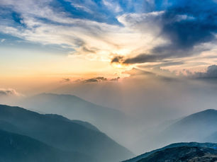 Ein atemberaubender Blick auf die Sonnenstrahlen, die in der Dämmerung durch die Wolken über nebligen Bergketten brechen. Der Himmel ist in Blau- und Orangetönen gehalten, während die Berge im Vordergrund Silhouetten bilden.