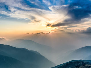 Een adembenemend uitzicht op zonnestralen die door de wolken breken boven mistige bergketens bij zonsopgang. De lucht is geschilderd met tinten blauw en oranje, terwijl de bergen gelaagde silhouetten op de voorgrond creëren.