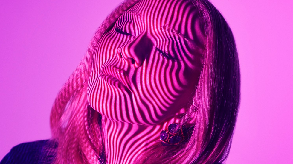 Close-up of a woman with eyes closed, her face striped by pink and purple light against a soft magenta studio background.