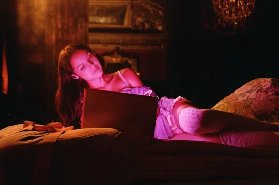 Woman lounging on a bed with a laptop in pink light, in a dark cozy room.