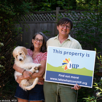 A man and woman stand in front of their garden. The woman holds a puppy and the man a sign that says: "This property is HIP"