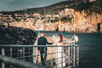 Bride and groom walking hand in hand away from the camera towards the Mediterranean Sea on a Sorrento Coast cliff.