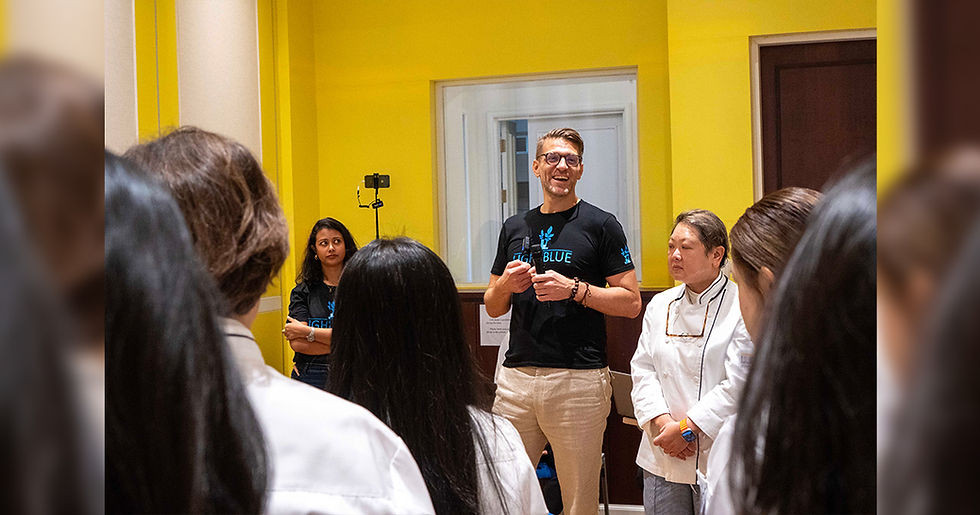 Man speaking to a small group in a yellow room, wearing a "Light Blue" shirt. Others look attentive. Mood is positive and engaged.