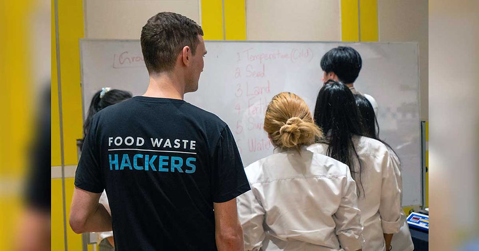 Four people face a whiteboard with handwriting. One wears a "Food Waste Hackers" shirt. Bright room, learning atmosphere.