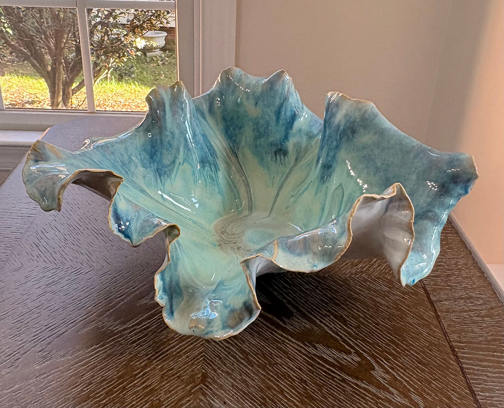 Ceramic bowl with ruffled edges, glazed in blue and green, sits on a wooden table by a window. Sunlight highlights its glossy surface.