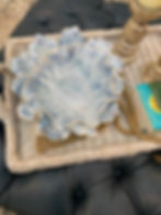 Blue ceramic bowl on woven tray with books and wooden beads, atop dark leather ottoman. Rustic candles in background. Cozy feel.