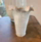 Elegant, white ceramic vase with ruffled rim on wooden table, large white vase and window in background, soft natural light.