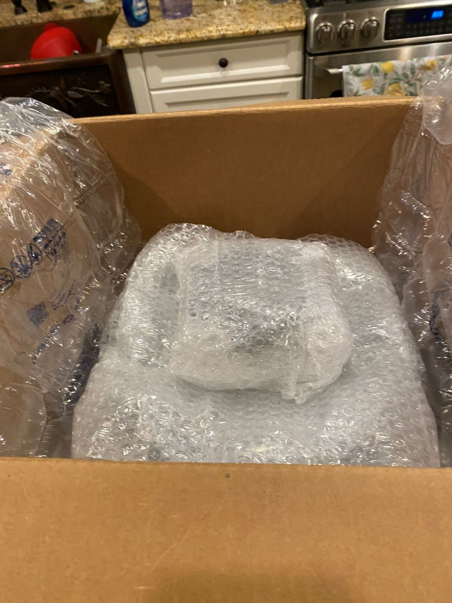 Cardboard box on kitchen counter with bubble-wrapped item inside. Air pillows on sides. Granite countertop and stove visible in background.