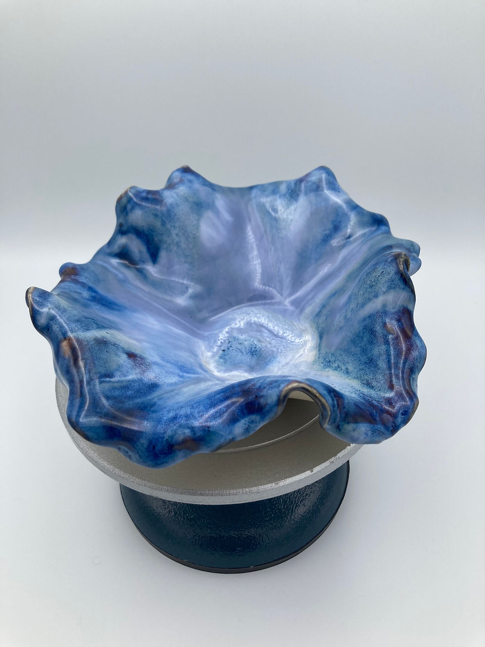 Thumbnail: Blue, wavy ceramic bowl on a round pedestal. The surface has a glossy finish with swirled shades of blue and white. Simple gr