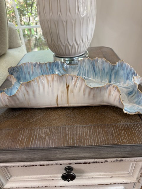 A wavy, ceramic platter with blue and brown tones sits on a wooden table, next to a textured white vase in a cozy room with soft lighting.