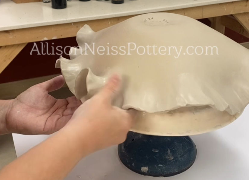 Hands shaping a clay piece on a blue pottery wheel. Background shows a workspace. 
