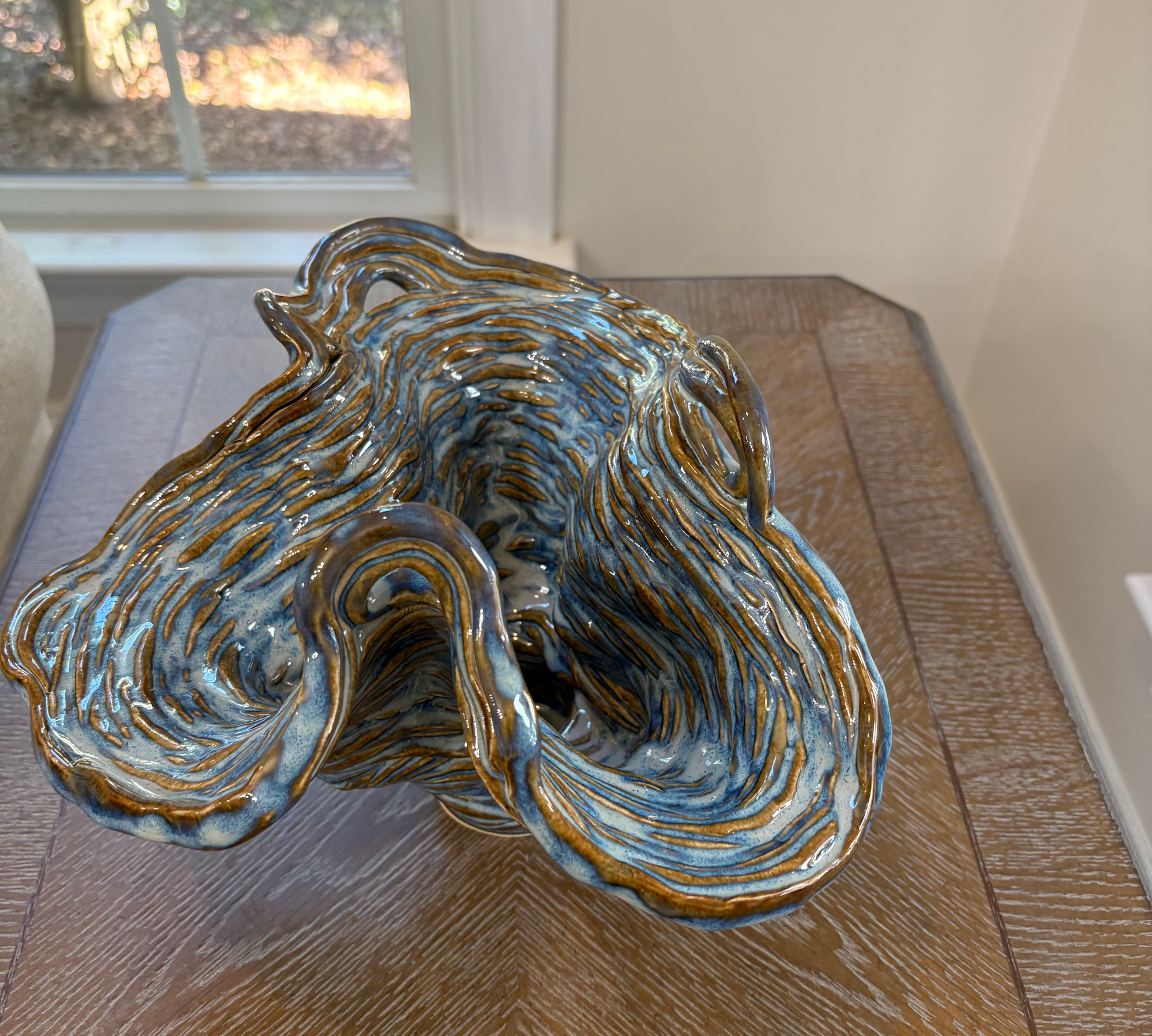 Glazed blue-and-brown ceramic sculpture on a wooden table by a sunlit window, with flowing organic folds and no text.