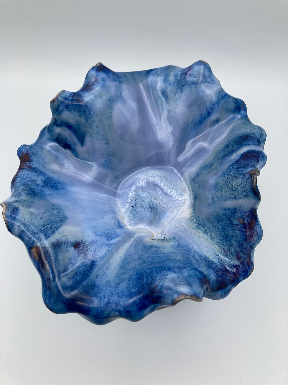 Thumbnail: Blue, wavy ceramic bowl on a round pedestal. The surface has a glossy finish with swirled shades of blue and white. Simple gr