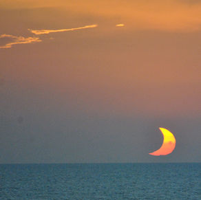 partially eclipsed sun setting low over a calm ocean under a dusky, warm-toned sky with soft clouds