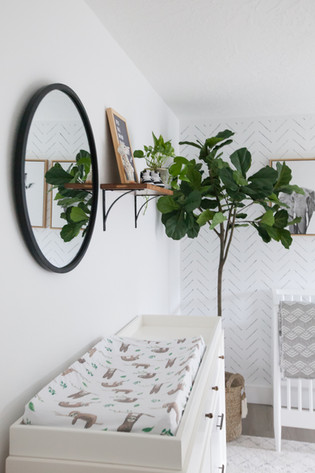 A bright and minimal baby nursery with white walls, a round black framed mirror, tall fiddle leaf fig, and white mid-century changing table. Room designed by Parlour & Palm of Portland, Oregon.