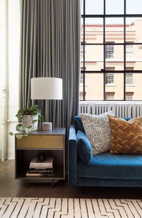 A light and airy loft style modern living room with tall ceilings, huge black-framed industrial windows with city views, a blue velvet sectional, rust velvet accent chairs, a geometric rug, modern coffee table and side tables. Designed by Parlour & Palm of Portland, Oregon.