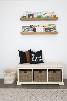The reading area of a bright and minimal baby nursery with white walls, wood picture ledges for displaying children’s books, a bench seat with storage baskets, a geometric rug, and a toy basket. Room designed by Parlour & Palm of Portland, Oregon.