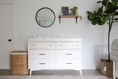 A bright and minimal baby nursery with white walls, a white mid-century modern dresser changing table, a geometric rug, round black framed wall mirror, wood shelf, tiny baby Nike shoes, a tall fiddle leaf fig in a basket, and a woven hamper. Room designed by Parlour & Palm of Portland, Oregon.