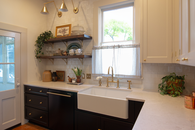 Two open wooden shelves with brass brackets in a kitchen styled with art, plants, and dishes. Creamy white tiles as a backsplash, White marble countertops. A fluted farmhouse sink with gold faucet.