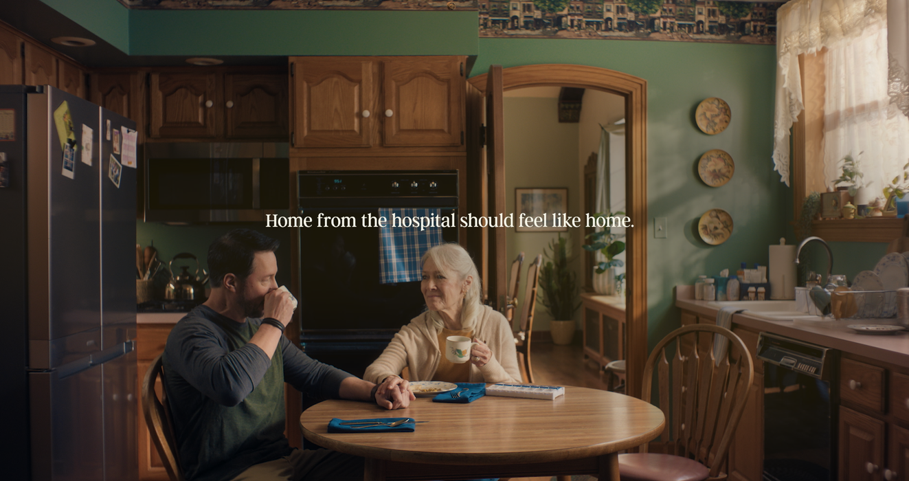 An older woman and her adult son sit at a round kitchen table holding hands and drinking coffee in a home hospice commercial scene.