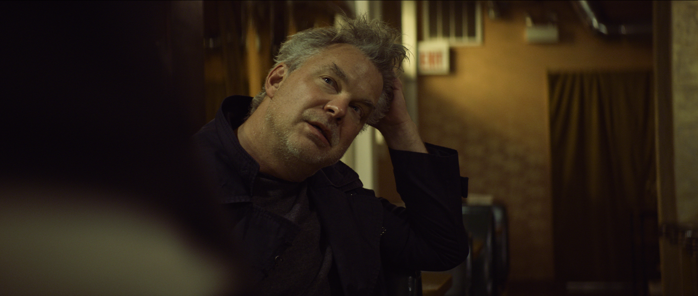 Peter DeFaria as Ronald leaning on his hand during a conversation in a dimly lit diner scene.