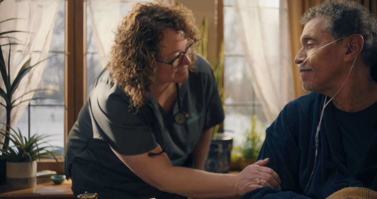 A nurse leans in to comfort and speak with an older male patient seated near a bright window in a home healthcare commercial. nurse with patient at home, Commonheart nurse visit, male patient healthcare ad, Robert Bennett, Missy Bille, home hospice scene, Chicago cinematography, Gabriel Jung