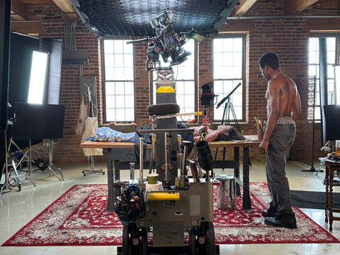 Cinema camera mounted on overhead rig filming an actor lying on a table inside an art studio set.