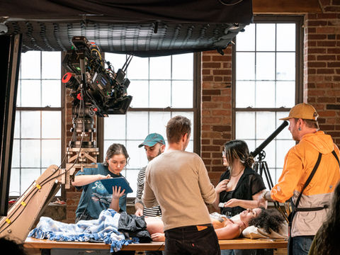 Film crew operating cinema camera while actor works at art table.
