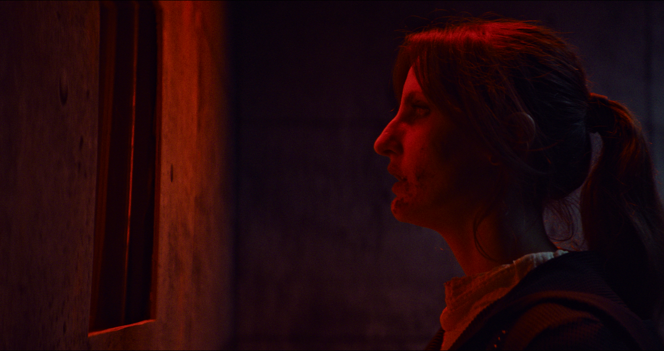 Woman in profile lit by red light inside a concrete chamber.