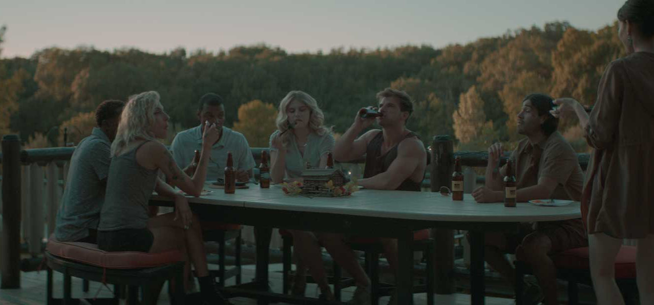 Group of friends sitting around a wooden outdoor table drinking beer on a cabin deck overlooking a forest and lake.