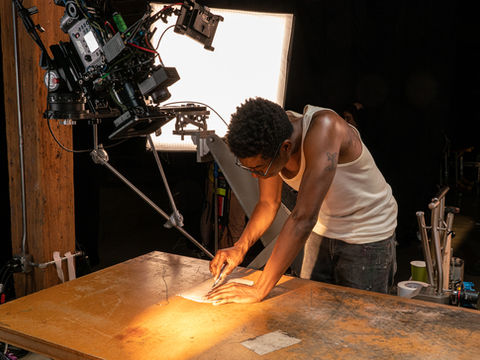 Film crew operating cinema camera while actor works at art table.