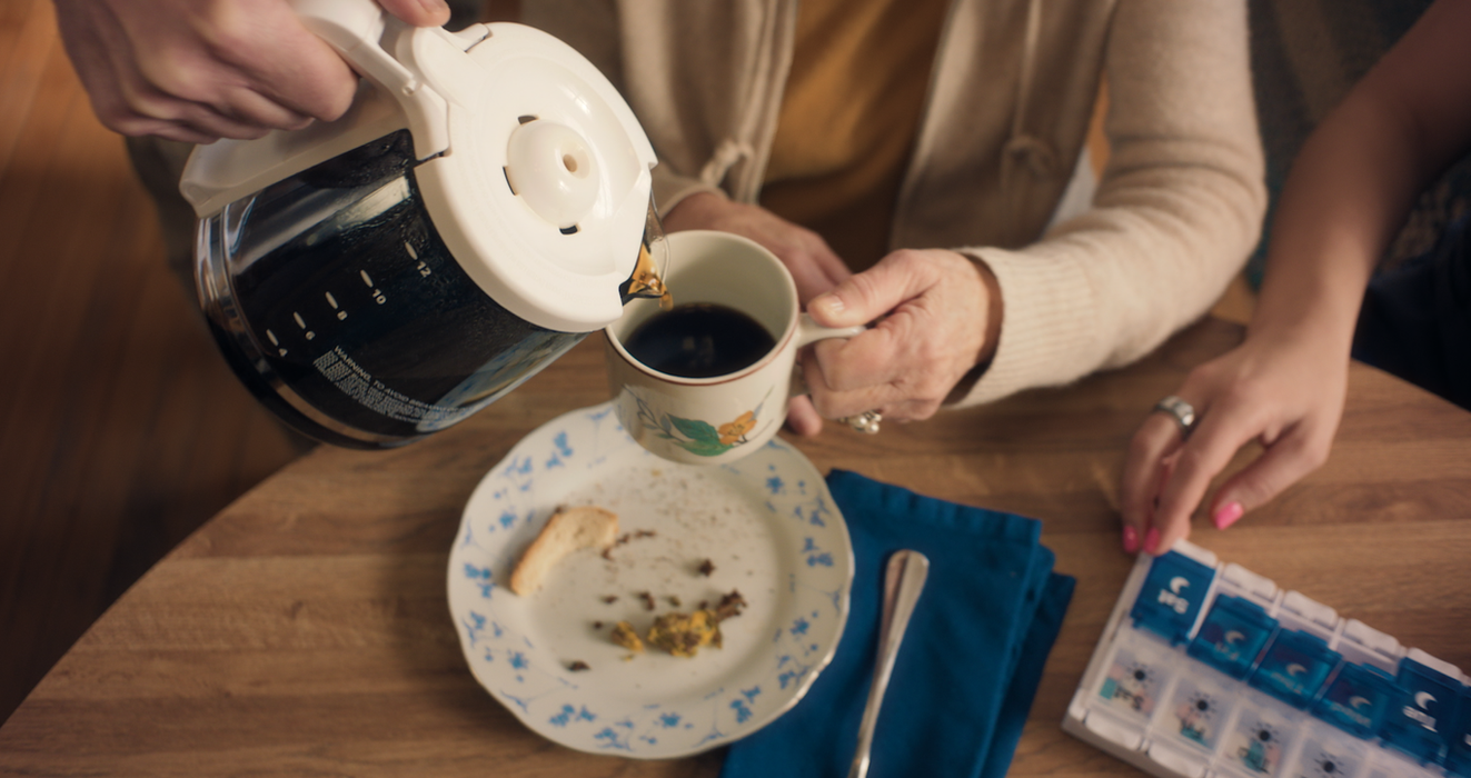 Close-up of coffee being poured into a floral mug at a wooden kitchen table beside a pill organizer, blue napkin, and breakfast plate in a Commonheart home healthcare commercial.
