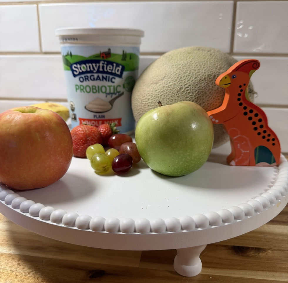 What you need for a volcano fruit bowl: yogurt, apples, strawberries, grapes, a banana and a cantaloupe, with a wooden toy dinosaur