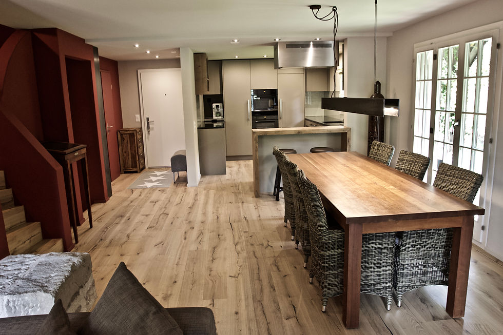 Das neue Wohnzimmer mit offener Küche, nach dem Umbau. Eine neue Wohlfühlzone in warmen Farben und Materialien. Die alten Bodenbeläge im ganzen Erdgeschoss und auf den Treppen werden entfernt und mit geöltem, lebhaftem Eichenparkett belegt. Im Bereich von Küche und Entrée wird die Decke abgehängt, um alle neuen Leitungen und die LED-Einbauleuchten unterzubringen. Wände und Decken werden gemäss neuem Farbkonzept mit Farbe aus Naturpigmenten gestrichen. Die neue, schlammfarbige Küche mit der kaffeebraunen Steinabdeckung und den speziellen Glasrückwänden, erhält schönes Zusammenspiel mit der niedrigen Bar und dem Spirituosenregal in matt lackiertem Eichenholz. Eingesetzte, edelstahlfarbig eloxierte Griffprofile in Kombination mit langen Stangengriffen in mattem Edelstahl passen perfekt ins Bild. Die dazu passende Küchenarmatur und die Dunstabzughutte in Sonderanfertigung sind ebenfalls in Edelstahl.