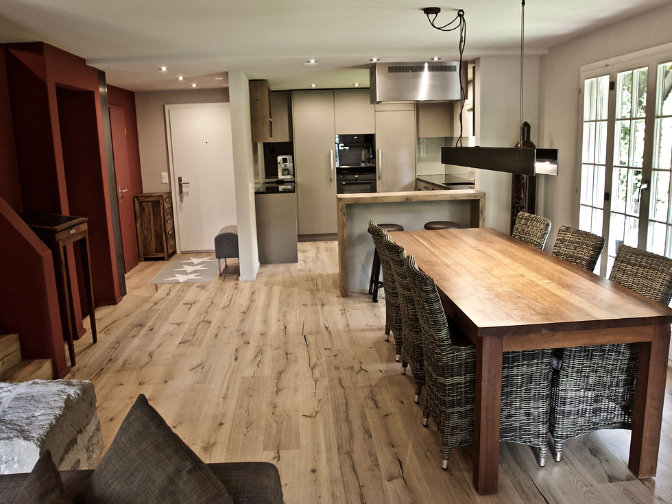 Das neue Wohnzimmer mit offener Küche, nach dem Umbau. Eine neue Wohlfühlzone in warmen Farben und Materialien. Die alten Bodenbeläge im ganzen Erdgeschoss und auf den Treppen werden entfernt und mit geöltem, lebhaftem Eichenparkett belegt. Im Bereich von Küche und Entrée wird die Decke abgehängt, um alle neuen Leitungen und die LED-Einbauleuchten unterzubringen. Wände und Decken werden gemäss neuem Farbkonzept mit Farbe aus Naturpigmenten gestrichen. Die neue, schlammfarbige Küche mit der kaffeebraunen Steinabdeckung und den speziellen Glasrückwänden, erhält schönes Zusammenspiel mit der niedrigen Bar und dem Spirituosenregal in matt lackiertem Eichenholz. Eingesetzte, edelstahlfarbig eloxierte Griffprofile in Kombination mit langen Stangengriffen in mattem Edelstahl passen perfekt ins Bild. Die dazu passende Küchenarmatur und die Dunstabzughutte in Sonderanfertigung sind ebenfalls in Edelstahl.