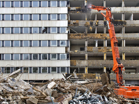 An orange excavator demolishes a building, revealing empty floors. Debris fills the foreground. The scene is industrial and dynamic.