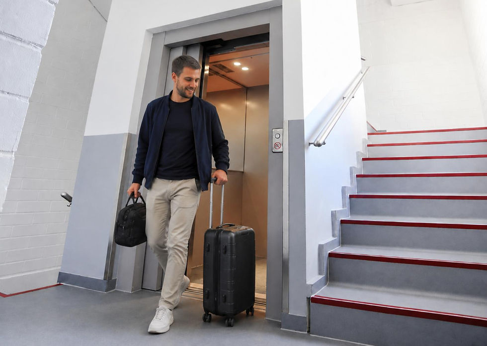 ein Mann kommt mit Koffer aus dem Aufzug gelaufen, rechts eine Treppe mit grauen Stufen und roten Kanten