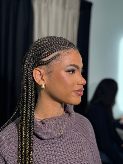 Soft glam makeup on a dark-skinned client with braided hair, featuring warm neutral eyeshadow, defined liner, and a glossy nude lip, photographed indoors.