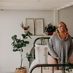 woman posing wearing long grey sweater on iron bed with greenery surrounding beige painted background