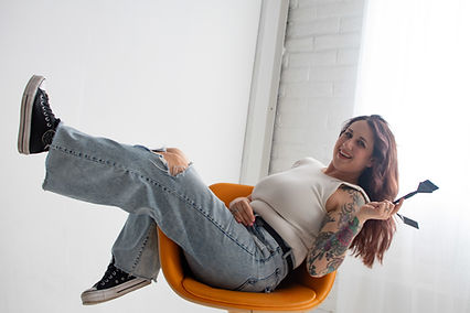 Relaxed woman with tattoos, holding paintbrushes in an orange chair.