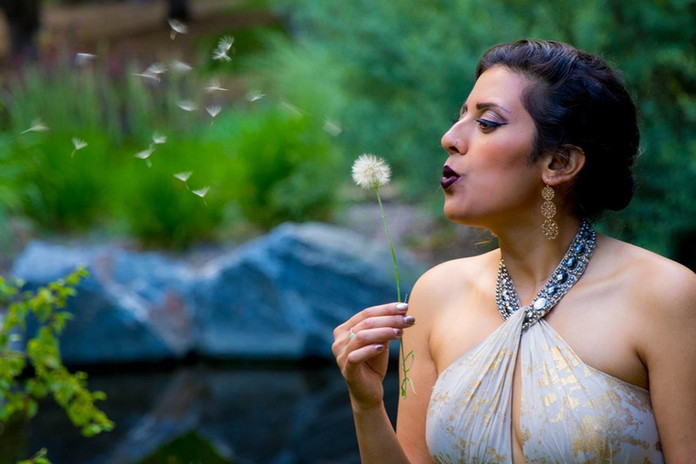 Woman blowing dandelion seeds, outdoors, with greenery in the background, EJS Beauty.